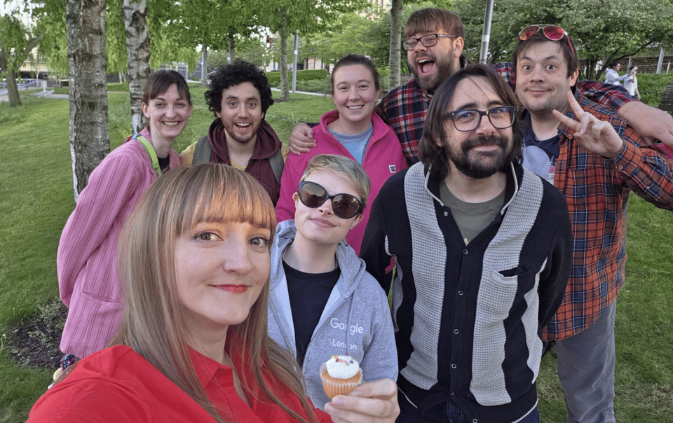 A group of eight smiling friends poses together outdoors, with one person holding a cupcake and others displaying various cheerful expressions.