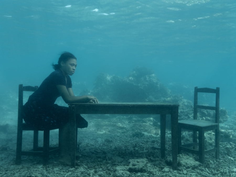 A girl sits at a table on the ocean floor, she looks into the distance, her hands resting on the table top.