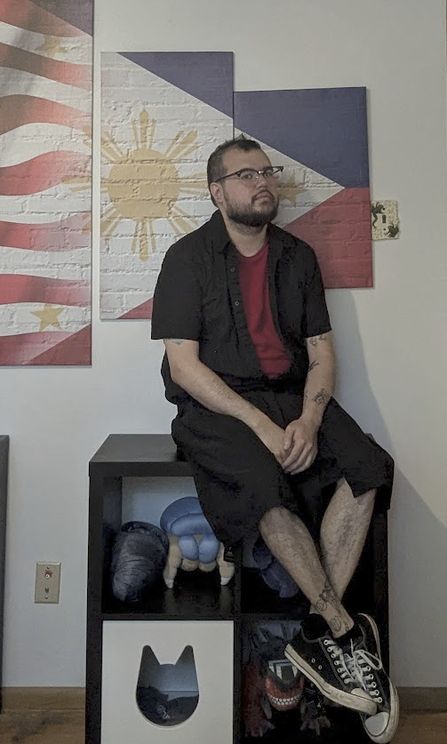 A picture of Mikkel sitting on a display case backed by a piece of art depicting the US flag transforming into the Phillipines Flag. They are wearing a black short sleeved collared shirt over a red shirt with a black shirt and black and white converses.