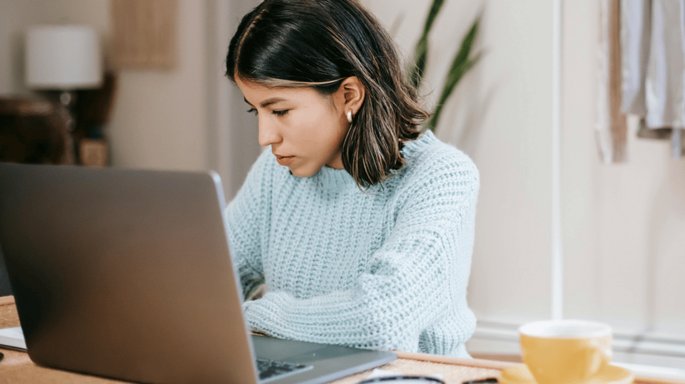 A lady wearing a sweater working on her computer with a mug on the side