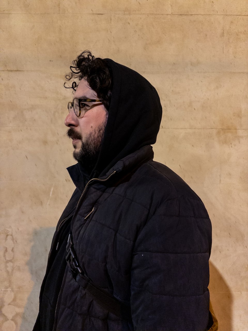 Jaime stands with his profile to the camera, wearing a black coat. His wet hair sticks out from underneath the hoodie.