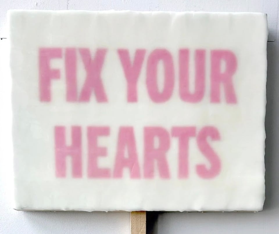 A wee painting that says FIX YOUR HEARTS in pink text on a white background. There's a stick at the bottom because it's a sign.