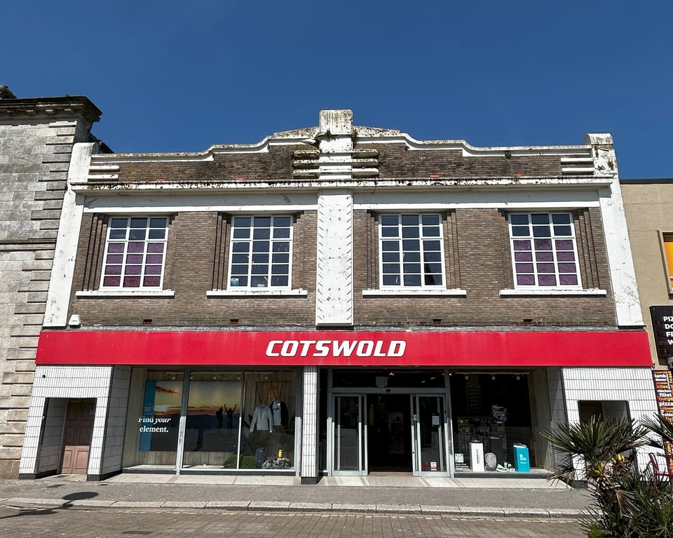 A simple two storey 1930s shop building with some lovely Deco details on the facade. There are four symmetrical windows on the first floor, each one holding 18 panes and set back slightly. Down the middle of the building is a wide white column with a zigzag bas relief on it. At the top are some decorative curved edges. Everything is in proportionAt the ground floor, there are two symmetrical side doors in tiled doorways. Then some idiot has removed the original windows and customer doors and fitted some aluminium frames that are very much not symmetrical.
