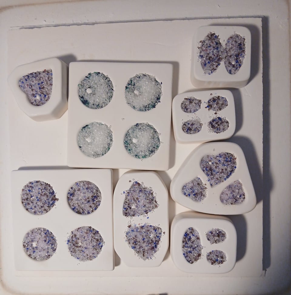 kiln shelf with various molds in a purple and blue and clear mix and some in a green blue mix. some of what's in there are strikers, meaning they won't come to color until heated, so there's gonna be some pink in there but there's not yet