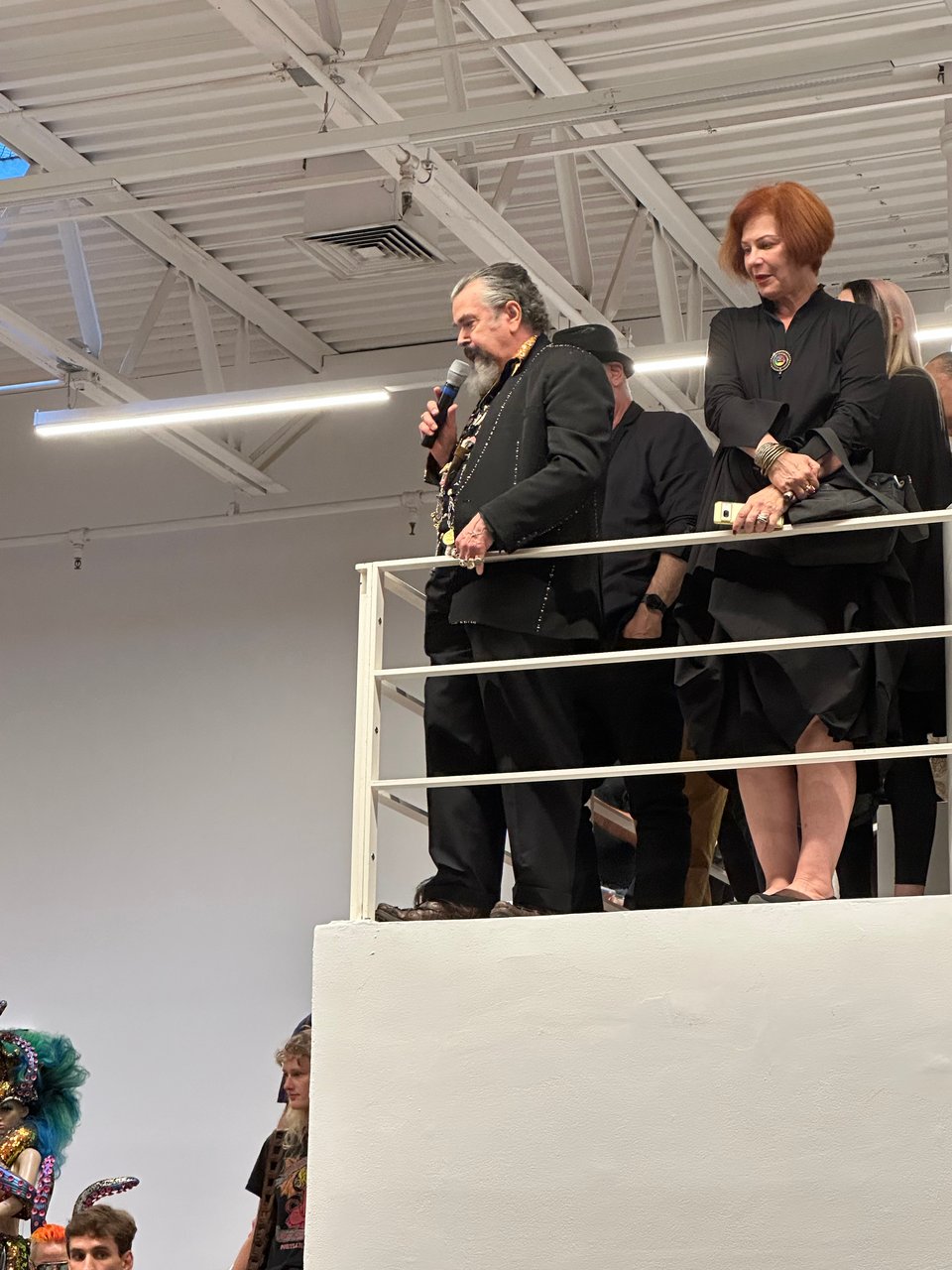 Photo of artist Joe Coleman on a raised platform at an art exhibit, holding a microphone and talking to the crowd