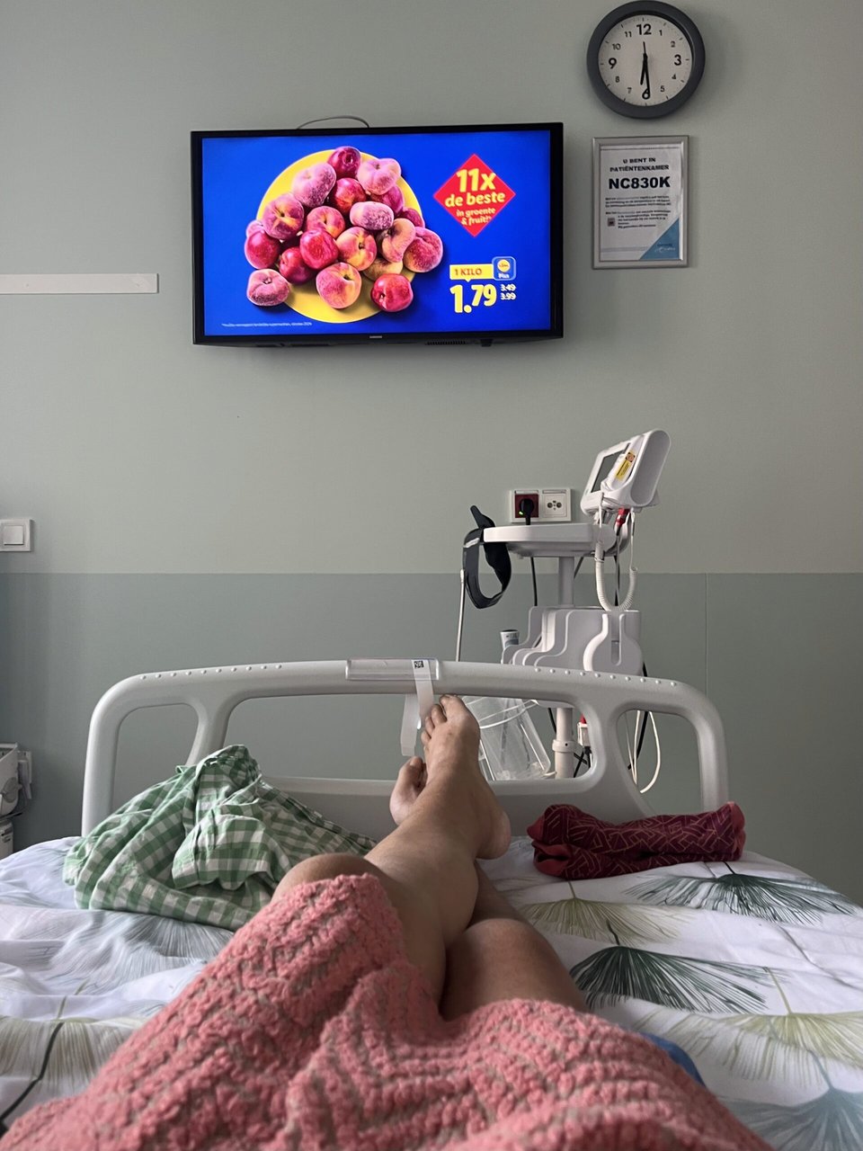 The perspective of the artist from the hospital bed; two legs outstretched with a view of the TV on a commercial for wild peaches at the grocery store Lidl.