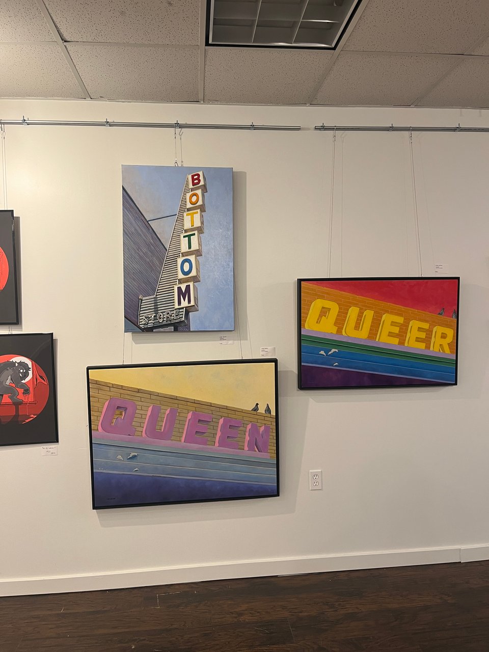 picture of an art gallery display with three paintings - one is a marquee sign with "BOTTOM" spelled out in rainbow letters and the others say "QUEEN" and "QUEER" against multicolored walls.