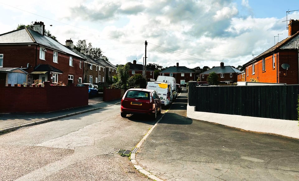 a colour photo of a cul-de-sac of semidetached houses. They are blocky and square, with a range of added on porches and satellite dishes. The gardens are mostly hidden by walls or wooden fences, and the road has vehicles parked along one side. There are no large trees beyond the houses.