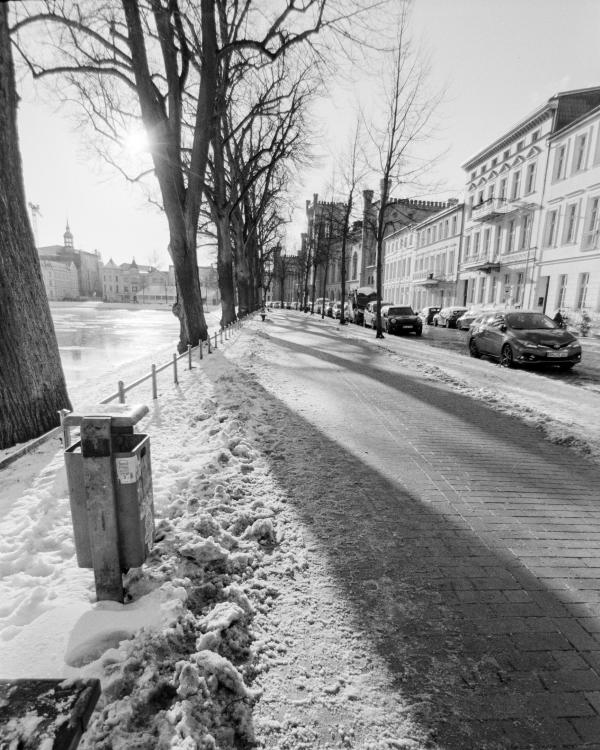B&W film photo of a snow-covered lakeside promenade in Schwerin with long shadows and historic buildings.
