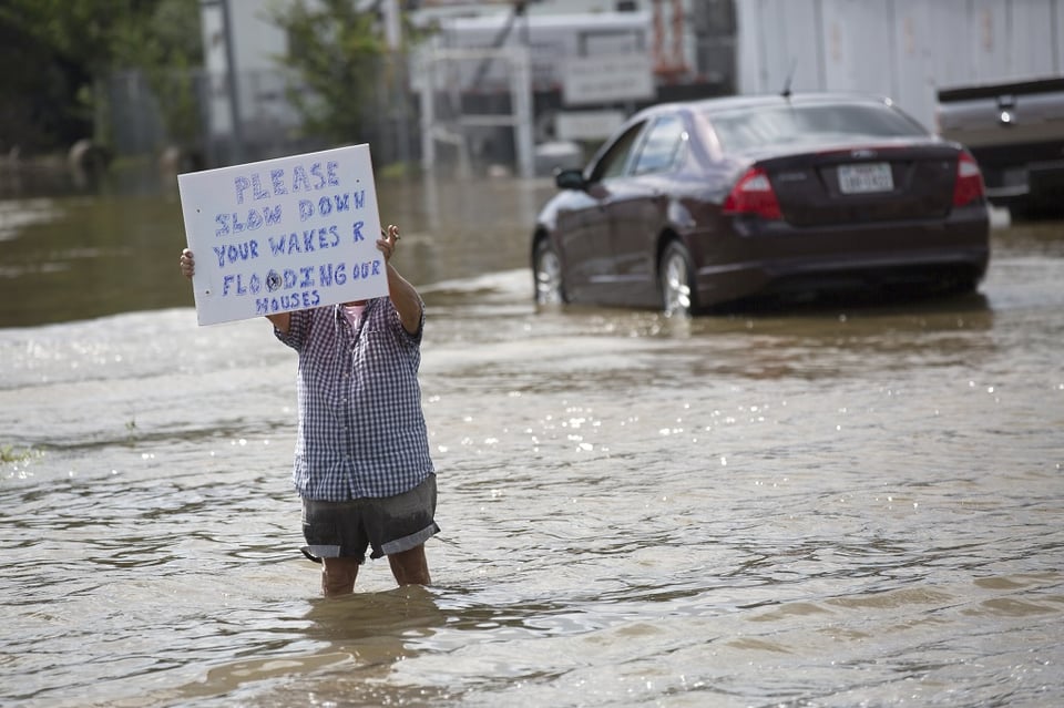 Someone standing in a flooded area, the water appears to go to their knees. They are holding up a homemade sign that says: “Please slow down your wakes R flooding our houses”.