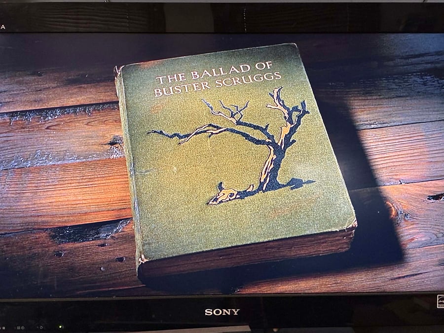 A photo of my TV, from when I was watching The Ballad of Buster Scruggs, specifically of when the title came on screen. This is a picture of an old book with a green cover on a hardwood floor. The book bears the title of the movie, and an image of a tree.