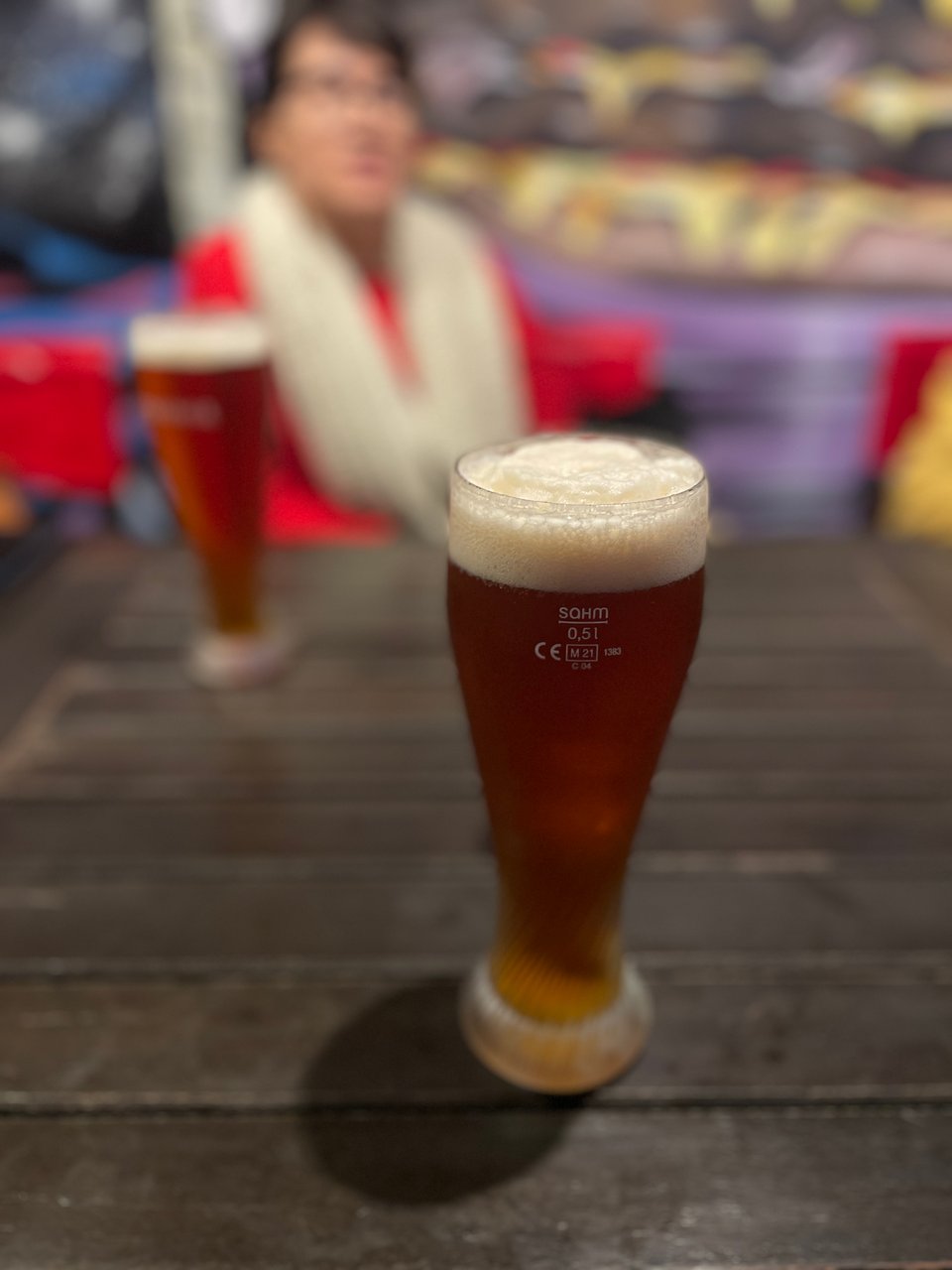 A tall glass of blond beer 1/2 liter is in focus in front. There is second glass in the background in front of a woman - both are blurred in the background.