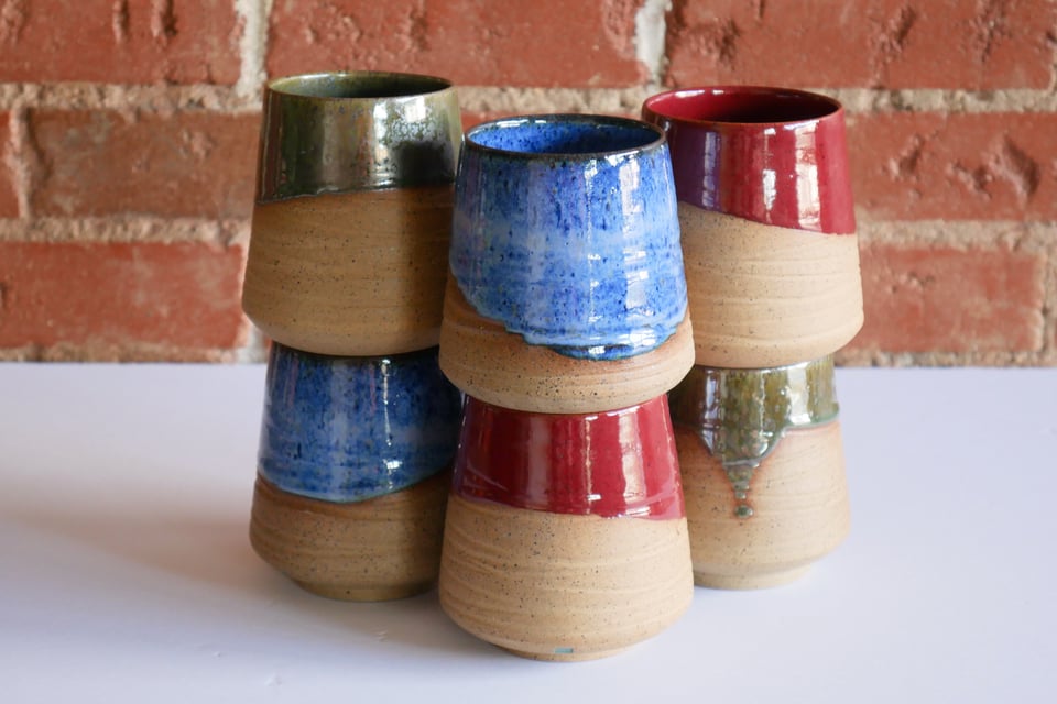 Six tumblers with a rounded base, textured specked inward-sloping sides, and colorful glazed rims in blue green and red