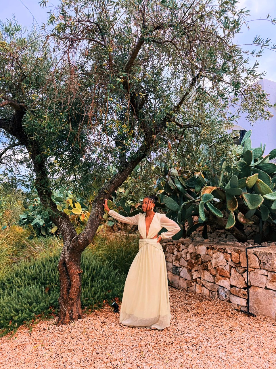 Nadia poses beneath an olive tree in a flowing light green dress, surrounded by greenery, cacti, and a stone wall in warm evening light.