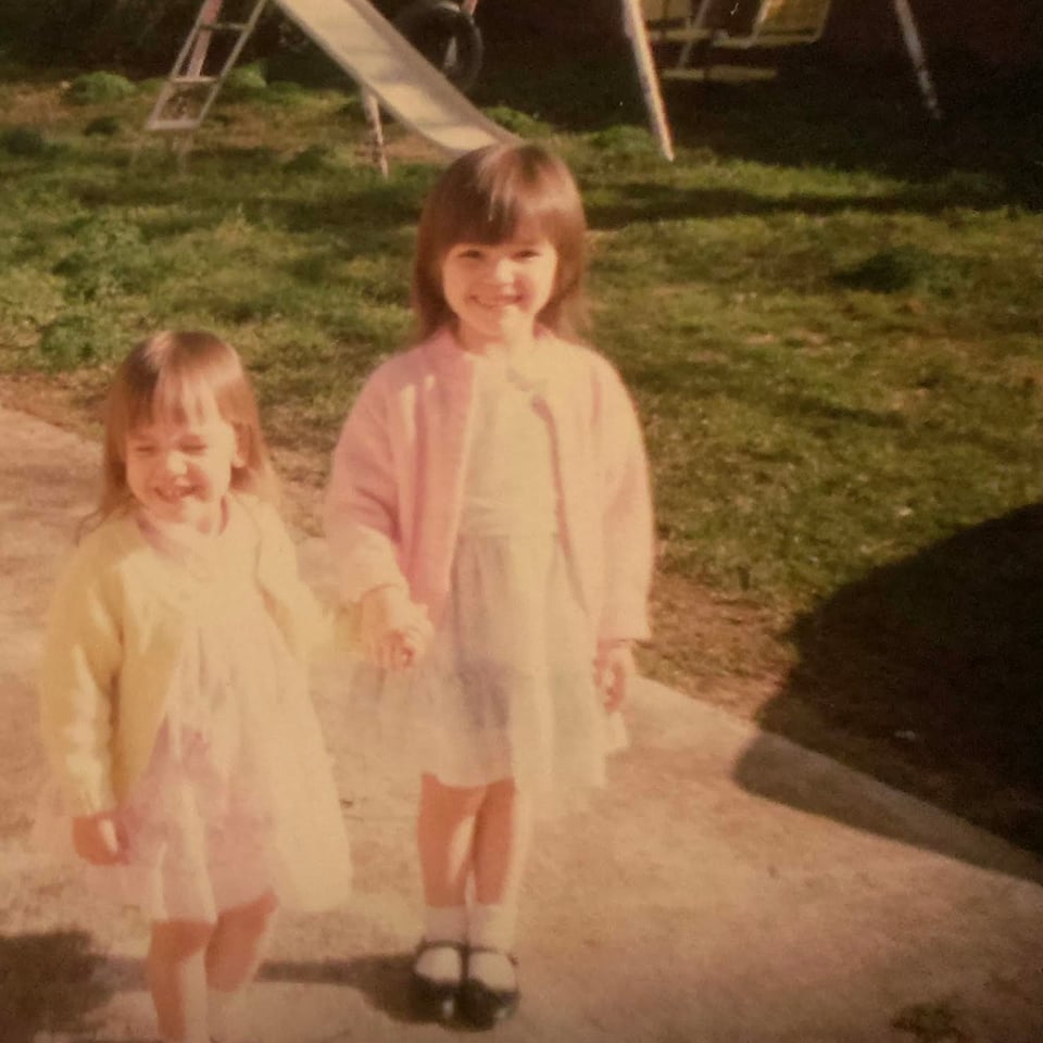 Two young brunette girls in pastel dressed smiling and holding hands in a yard