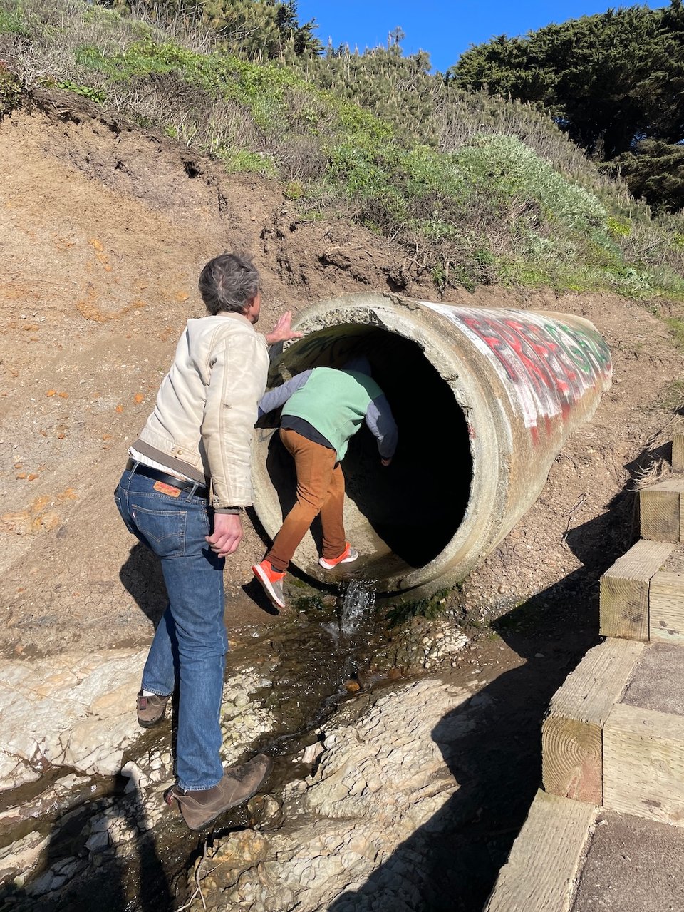 A young boy climbs through an oversized storm drain, his uncle waits behind him to enter the tunnel.