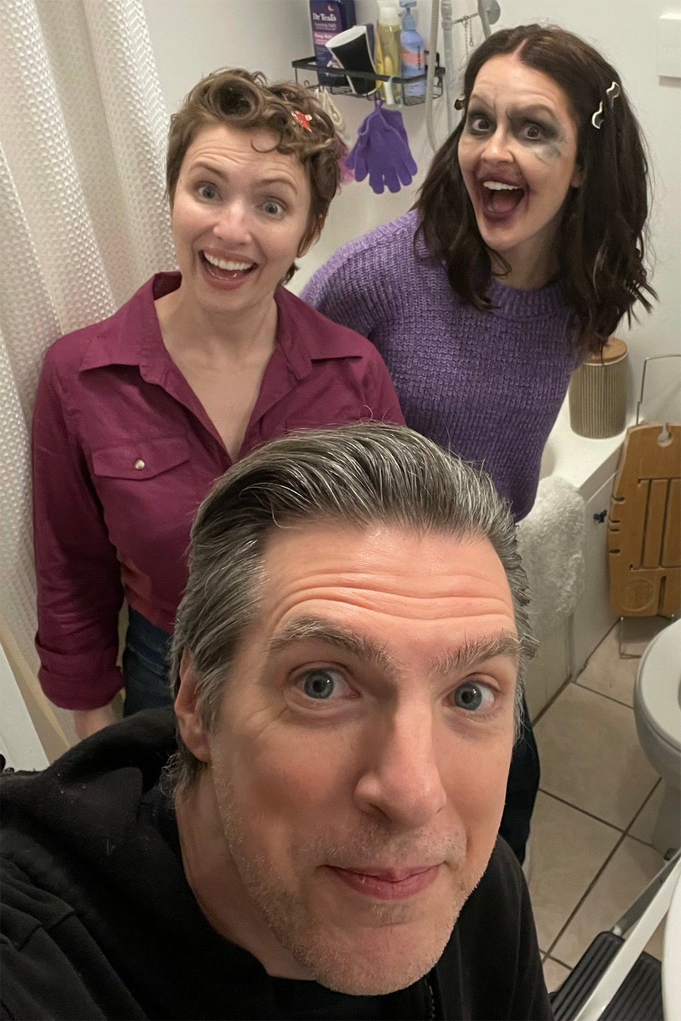 Three actors smiling at the camera in a bathroom