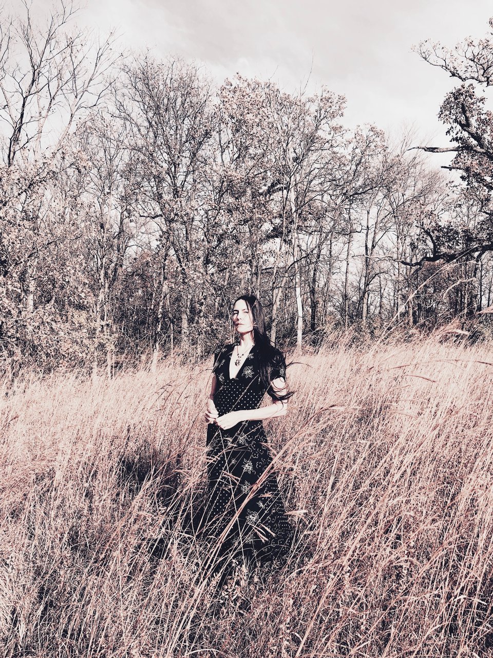 Jesse, a woman with long black hair and light skin, stands in a sunny field wearing a black print dress.