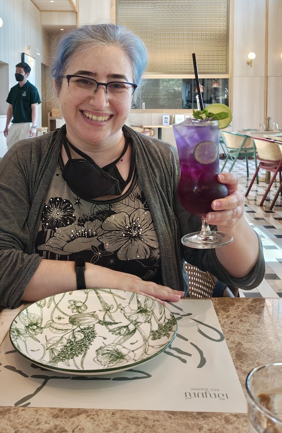 Me, a white woman with blue-purple gray hair sitting at a table at a very nice Thai restaurant, holding up a butterfly pea limeade, a purple drink in a hurricane glass garnished with pea shoots and lime wheels. The restaurant is done in light stone and off-white tones. The plate in front of me (while sadly empty) has a very nice botanical design in green on a white background.