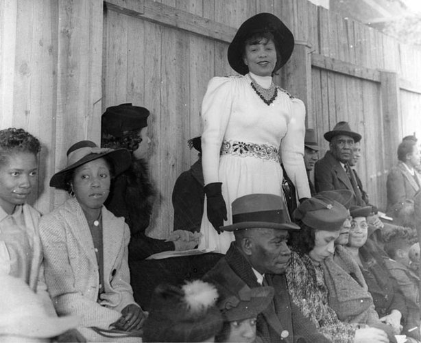 zora neale hurston standing in a crowd of seated black people. she wears a white belted dress and black hat and gloves.
