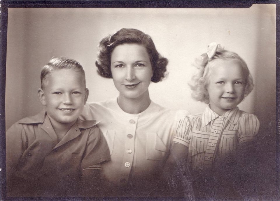 A photo of a cleancut white family in the 1940s or 1950s: boy with wide lapels, woman with a cute side-part bob and white blouse, young girl with blonde hair and a stripey shirt.