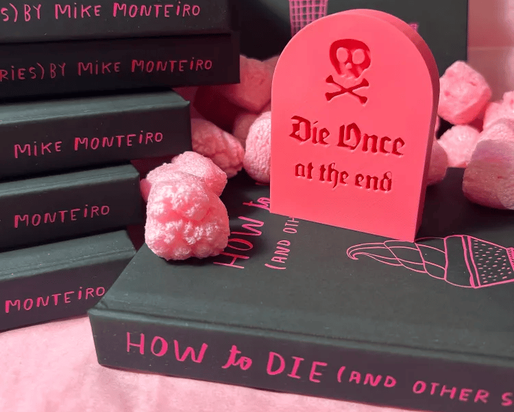 A little pink 3-d printed tombstone reading Die Once at the end, sitting on top of a copy of How to Die and other stories