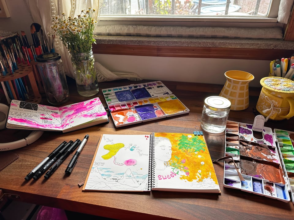 Jessi's studio in the daytime. It shows a slightly chaotic scene with paint supplies and multiple sketchbooks out alongside pens, water containers, a jar with chamomile flowers, a mug wrapped tight with a cozy and hat, and various art supplies. It's messy, but it's interesting.