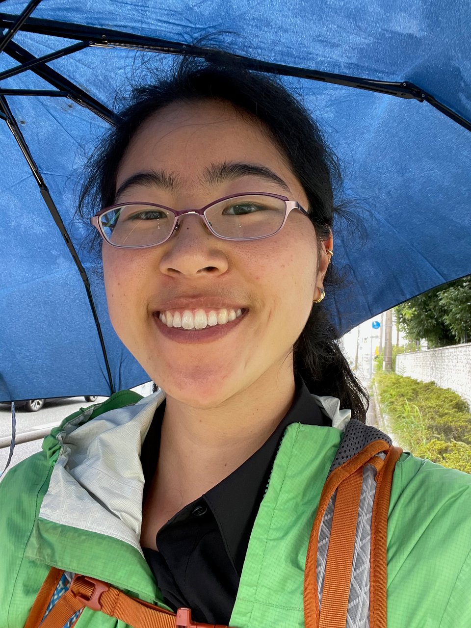 Selfie of the author wearing full rain regalia over a business appropriate outfit under the shelter of an umbrella. The author is smiling but inside they are cursing the rain.
