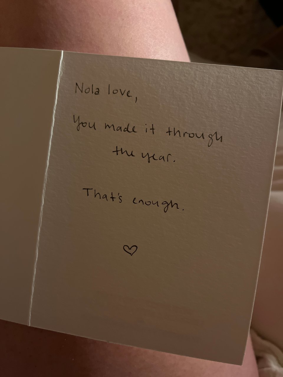 a handwritten card against bare skin, reading "Nola love, you made it through the year. That's enough." with a small heart