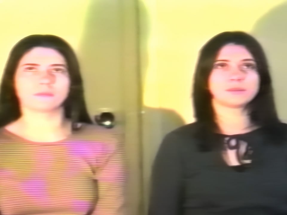 Two young white women with dark hair are shown sitting side-by-side in front of a yellow wall (identical twin artists Jane and Louise Wilson). The image is a still from grainy video footage shot in a motel room.
