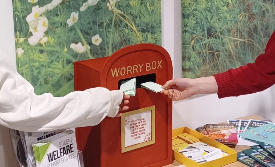 Two arms place folded notes into a small box shaped like a red British postal box, which says "Worry Box" on it