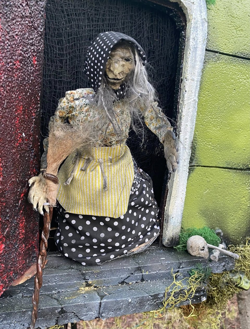 A close up of a sculpted doll of an old lady wearing a headscarf and apron, standing in the doorway of the hut. There are some bones nearby