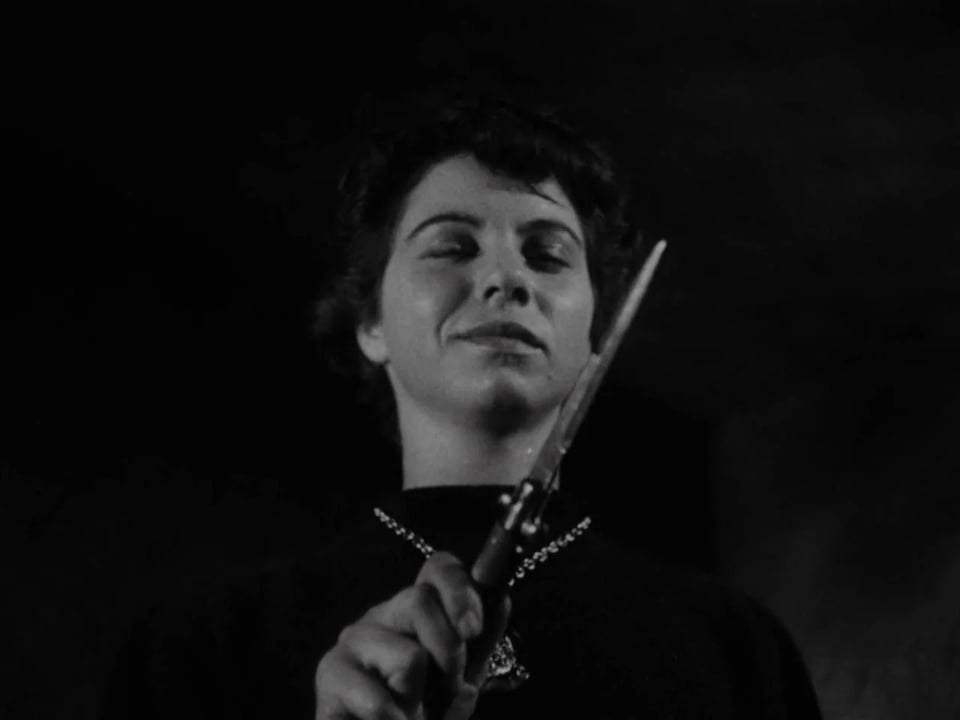 A smiling woman with short dark hair and a dark sweater contemplates the switchblade she holds in front of her.