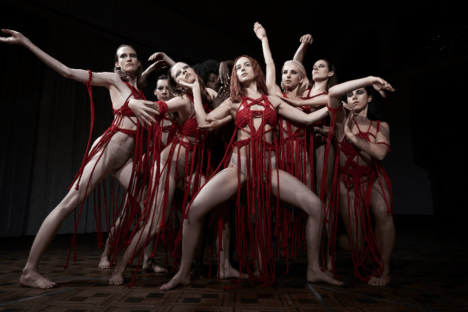 Seven dancers wearing very little except for red ribbons strike an expressionistic pose in Suspiria 2018.