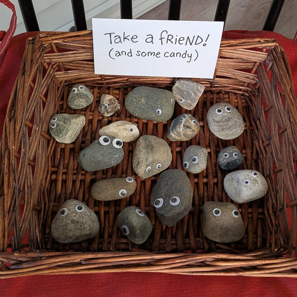 a square woven basket is filled with a small collection of smooth river rocks, each of which has been given a pair of googley eyes so they appear to be staring up at you. A handwritten sign in the basket reads "take a friend! (and some candy)"