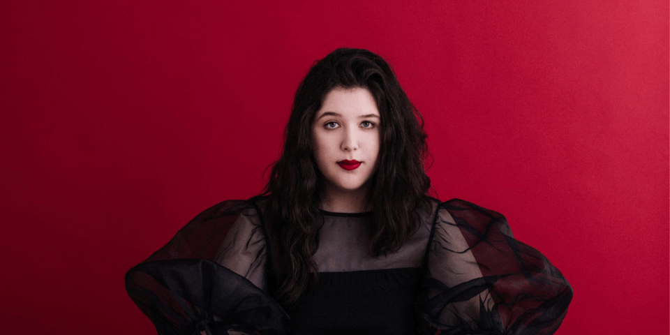 Lucy Dacus in front of a deep red background wearing a big-sleeved dress.