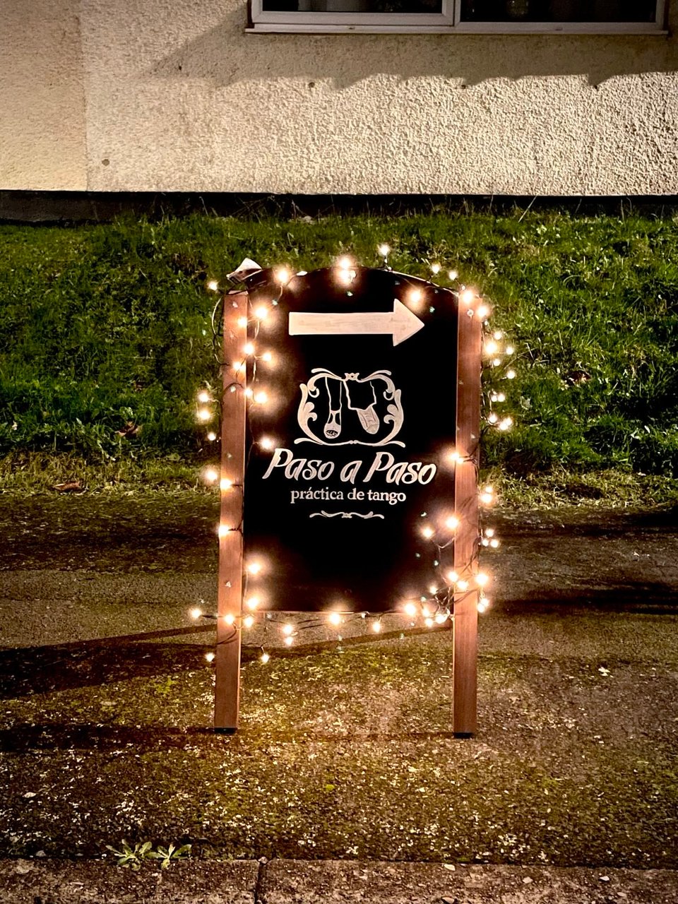 Blackboard pavement sign of Paso a Paso tango with fairy lights.