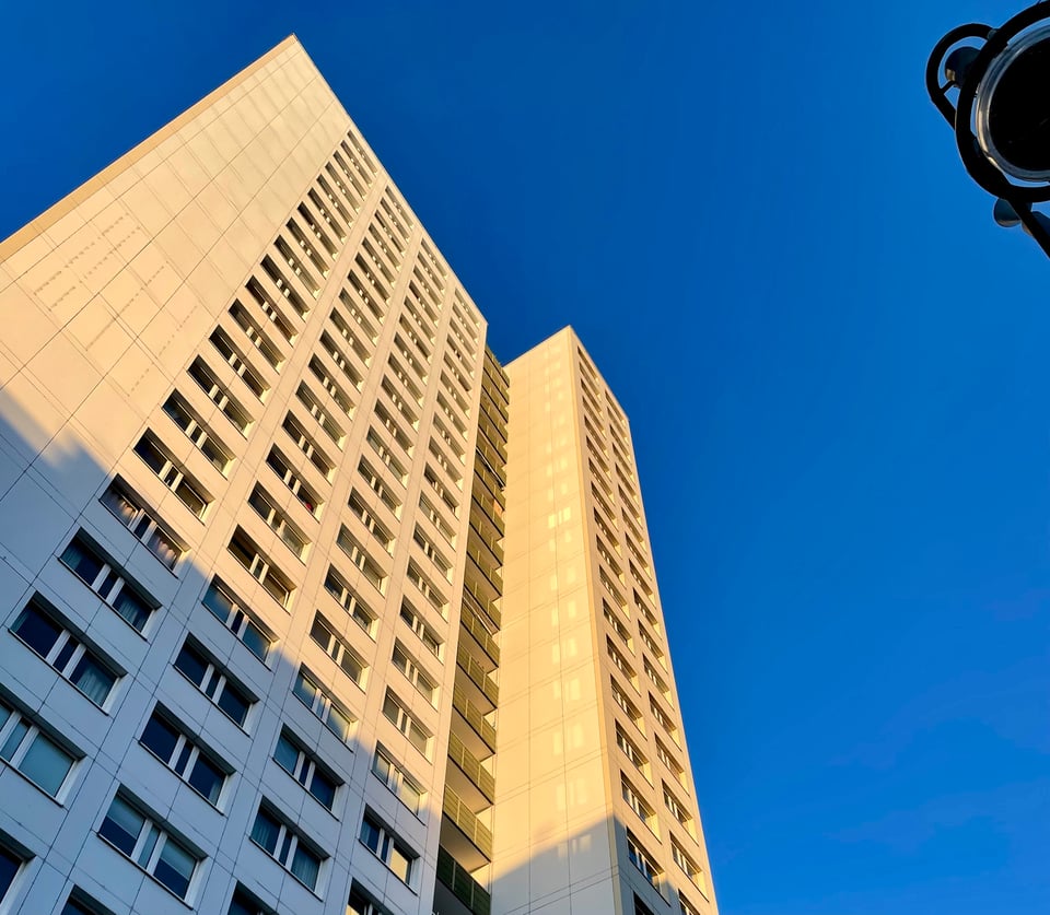 A view of a tall east german style building with the lower part covered in shadows due to the low sun (which is not visible in the photo) and upper part illuminated yellowish for the same reason. There is blank blue sky behind.