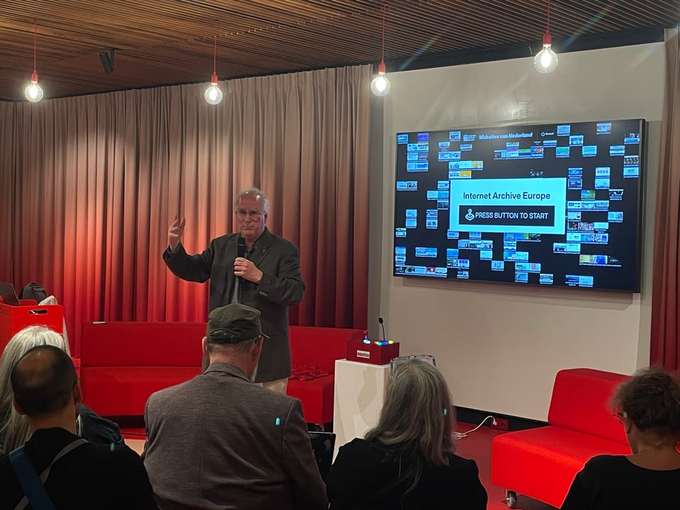 A man presents in front of an audience. The web archiving display is on a screen behind him.