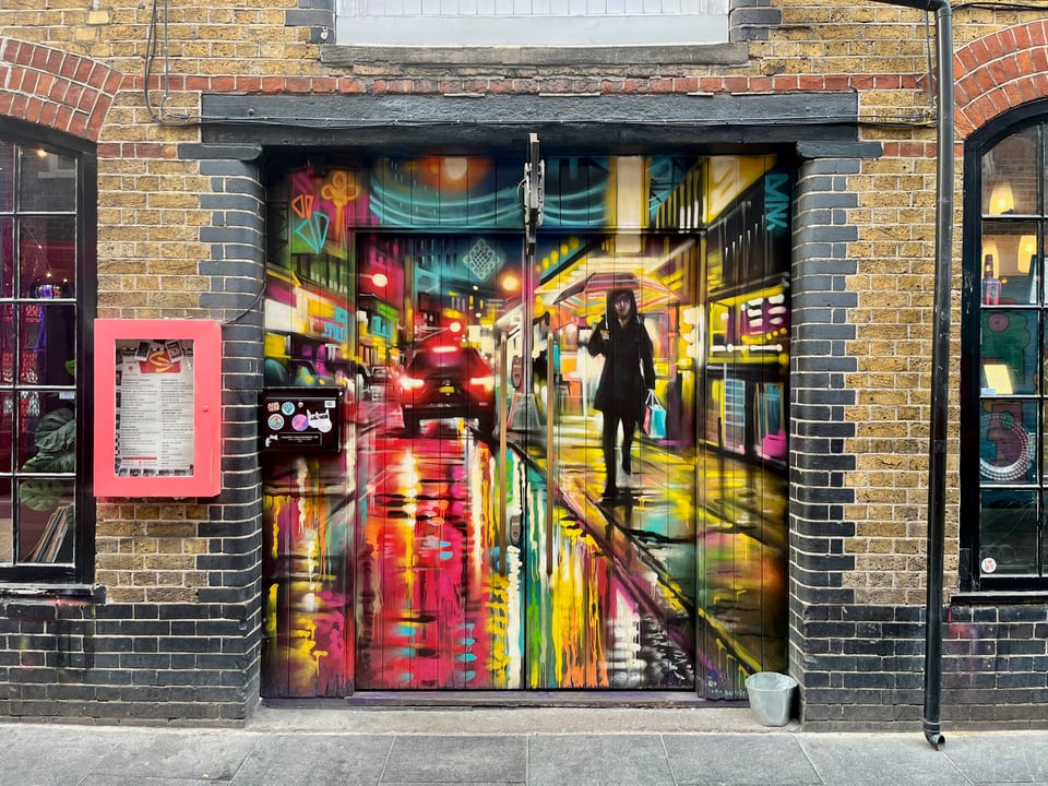 A colourful mural showing a city road at night, with bright lights in different colours. A person is walking towards the viewer along the pavement, holding an umbrella – it’s raining, and we can see colourful reflections in the road surface. The mural is in the middle of a plain brick wall, with windows on either side.