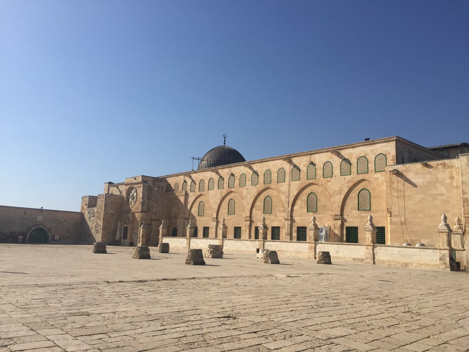 A three-story stone building with a small black dome made of sand-colored stone, in front of a plaza made of stone in the same color