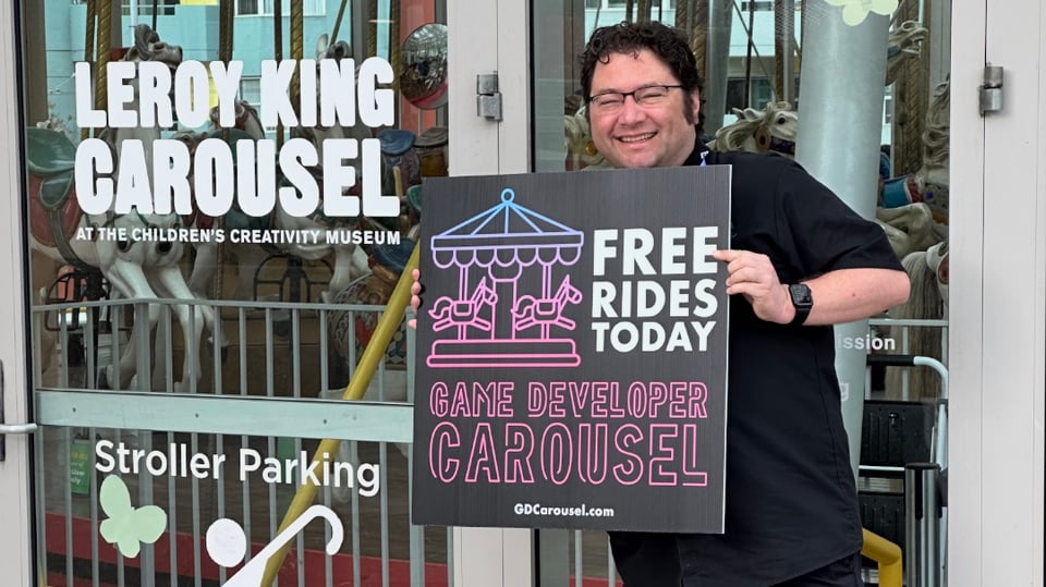 Seg showing off the Game Developer Carousel sign in front of the entrance to the LeRoy King Carousel.