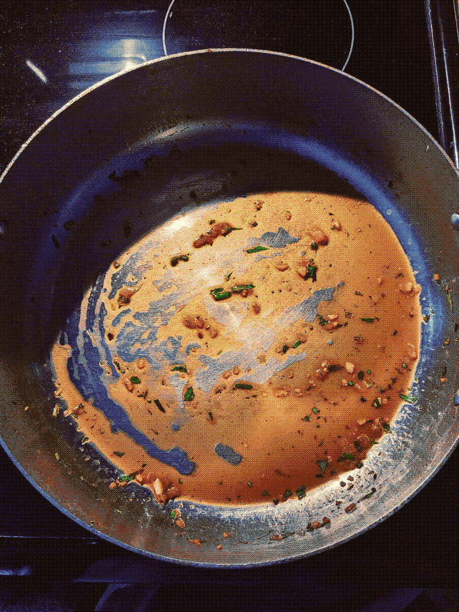 Top-down view of a high-walled steel pan. It is empty, but with a thin layer of brown emulsion and a few scattered herbs remaining on the bottom.
