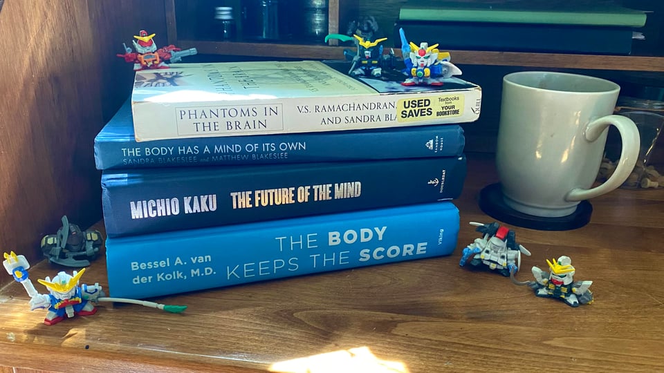 A stack of books: Phantoms in the Brain, The Body Has a Mind of it's Own, The Future of the Mind, and The Body Keeps the Score, on a wooden desk, with mini mecha figures on it, and a grey mug to the right.