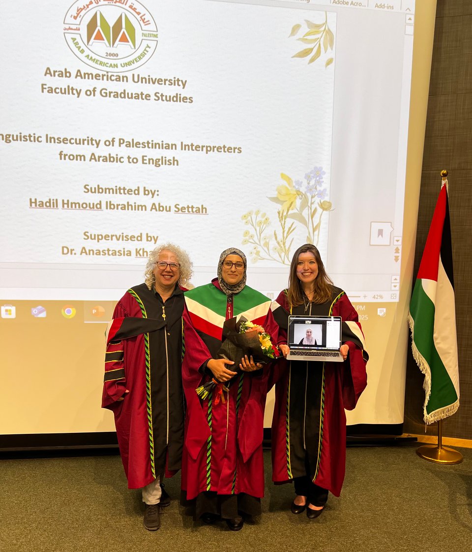 Three women standing in red academic regalia; the woman on the right holds a laptop with an image of a fourth woman via videoconference