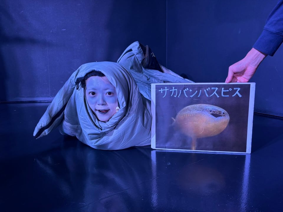 A person lies on the floor wrapped in a grey blanket. Someone holds up a picture of a specific blobby grey fish next to them
