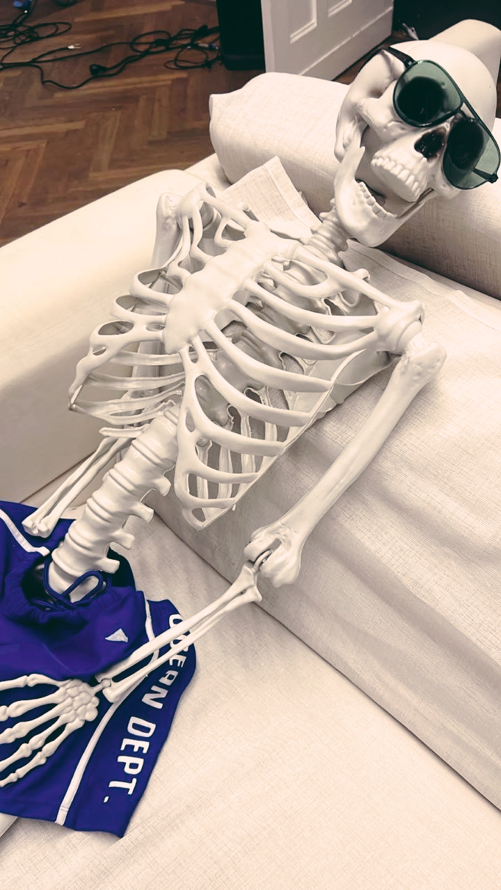 A photo of a skeleton wearing shorts and sun glasses sitting on a sofa
