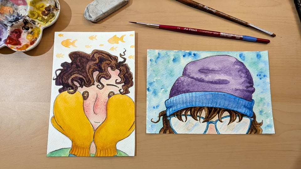 two small watercolor paintings on a desk. One features Iris, who has short curly dark brown hair and freckles, with sunny yellow mittens covering most of their face. The other is the top half of Vi's face, just glasses, brown bangs, and a slouchy purple and turquoise beanie.