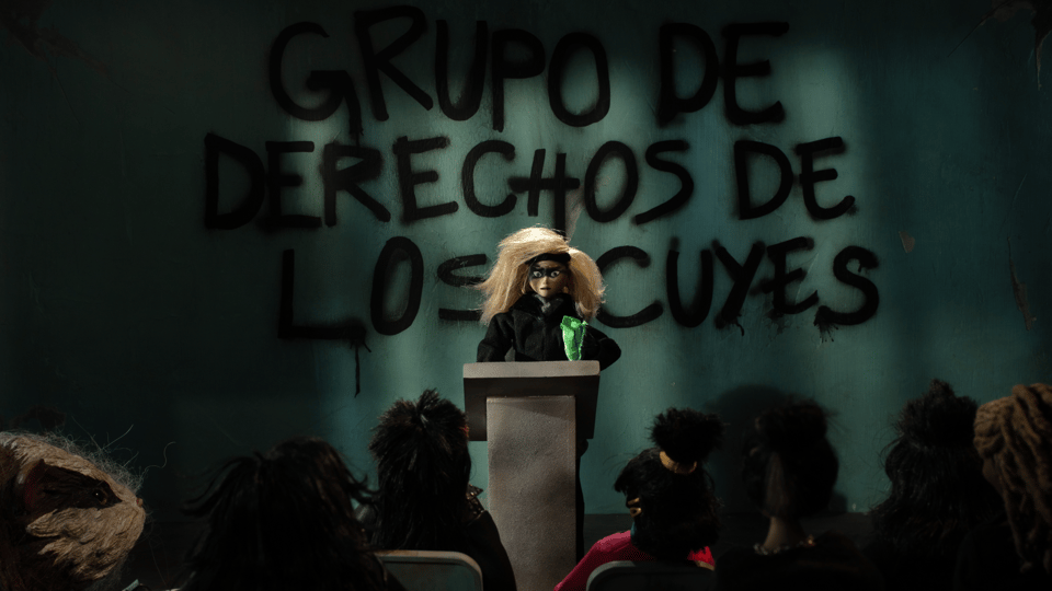 A darkened room, a woman stands in front of a podium, graffiti on the wall behind her