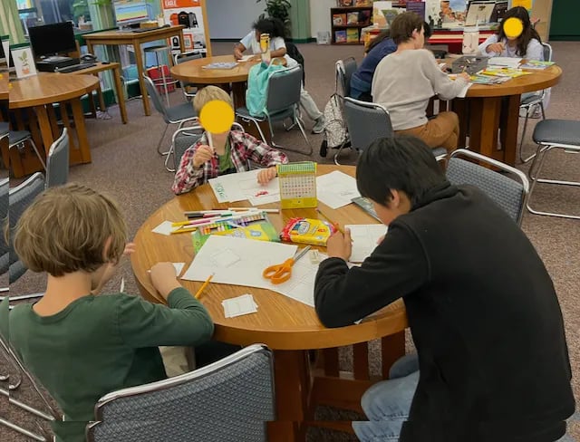 Image from an elementary school library. There are three round tables each with two or three elementary-aged students sitting around them, with comic books and drawing supplies scattered around.